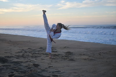 Woman practicing karate on beach against sky during sunset