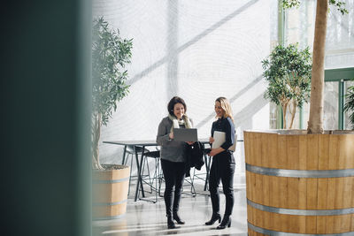 Businesswoman showing laptop to colleague while discussing in office corridor