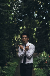 Portrait of young man gesturing while standing on footpath against trees