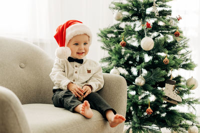 A cheerful boy in a santa claus hat is sitting under a christmas tree in an armchair 
