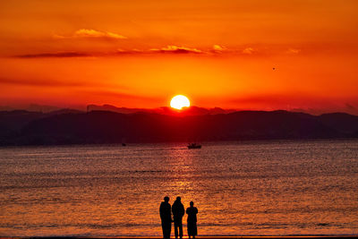 Silhouette people on shore against orange sky