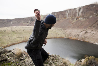 Side view of hiker throwing stone in lake while standing on cliff