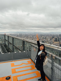 Portrait of woman with hand raised standing on building terrace against cloudy sky