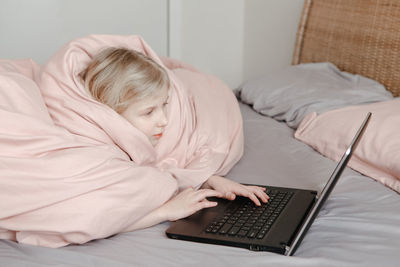 Bored girl lying in bed under blanket and learning in online school class. kid working on laptop