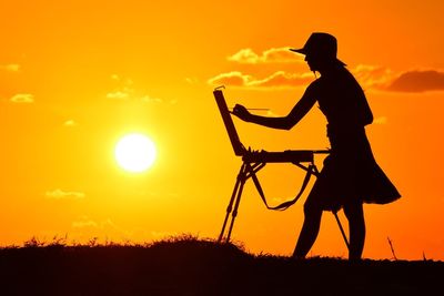 Silhouette woman painting while standing on field against orange sky