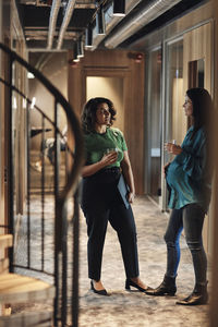 Businesswomen communicating with each other while having drink in corridor at office