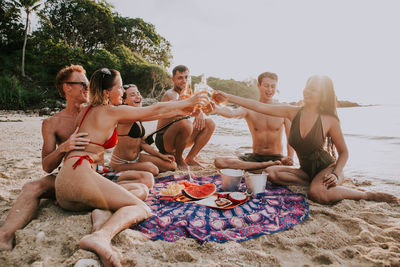 Friends toasting beer bottles while sitting at beach