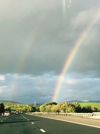 Scenic view of rainbow over country road against cloudy sky