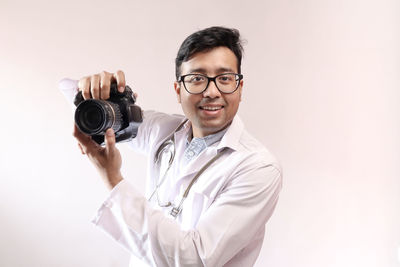 Portrait of smiling young man holding camera against white background