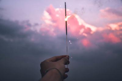 Cropped hand holding lit sparkler against sky during sunset