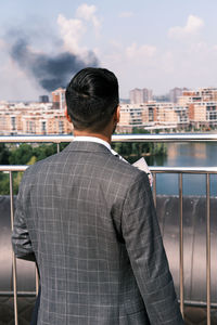Rear view of man standing by railing against buildings in city