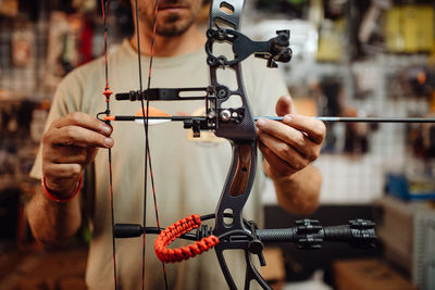 Cropped unrecognizable busy male hunter adjusting compound bow with arrow while standing in garage and preparing for shooting