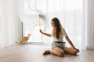 Caucasian teen girl sitting on the floor playing with a cat cornish rex with a stick teaser