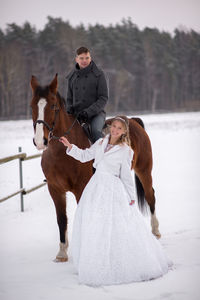 Portrait of smiling bride standing by groom sitting on horse at snow covered field