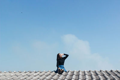 Woman shielding eyes while sitting on rooftop against sky