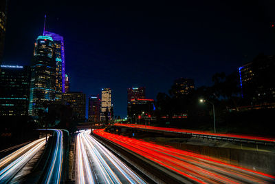 Light trails on road by illuminated buildings against sky at night