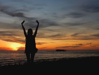 Silhouette woman with hand raised standing at beach against sky during sunset