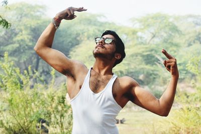 Young man gesturing while wearing sunglasses against trees