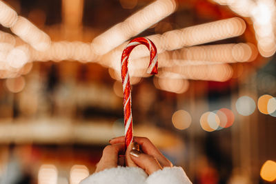 Female hands holding red white cane sweet lollipop against the background of golden garland lights