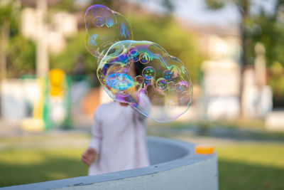 Obscured face of boy blowing bubbles while standing at park during sunny day