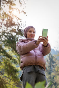 Low angle portrait of smiling man holding camera while standing against sky