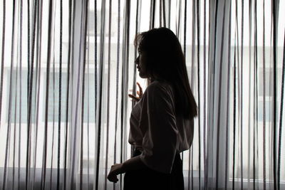 Side view of young woman looking through window at home