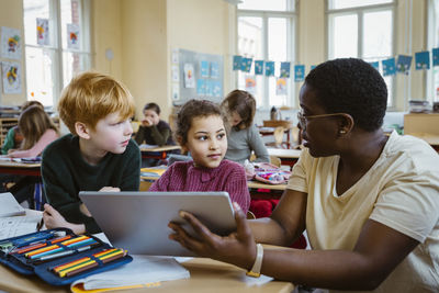 Female teacher assisting schoolboy and schoolgirl with digital tablet in classroom