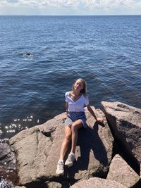 Rear view of woman sitting on rock by sea against sky