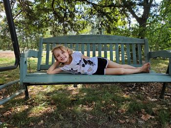 Portrait of smiling girl lying on bench against trees