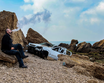 Man smoking while sitting on rock against sky