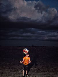 Boy playing with ball on field against cloudy sky