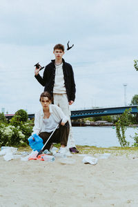 Full length portrait of confident young friends collecting plastic waste near river against sky