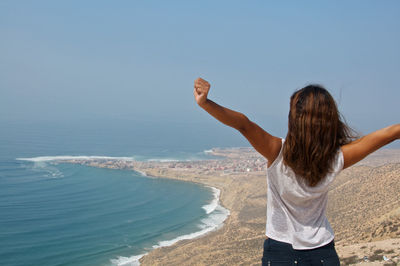 Rear view of woman with arms raised standing by beach against sky