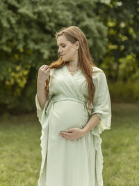 White pregnant woman holds and looks at strand of hair,puts hand on belly, standing in park