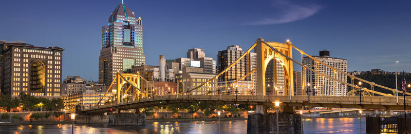 Illuminated bridge over river by buildings against sky in city