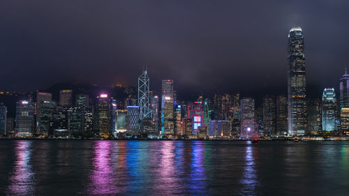 Illuminated buildings by river against sky at night