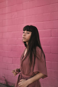 Side view of stylish young ethnic female with long dark hair in trendy dress standing against pink wall on street and looking away thoughtfully