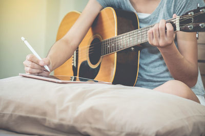 Midsection of woman playing guitar while using digital tablet on bed