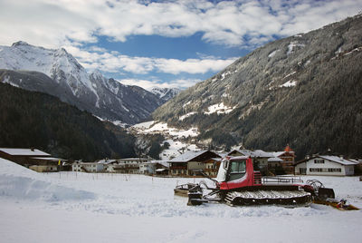 A view of the snowy mountains from the valley, with houses and a red snowcat in front