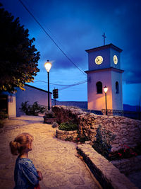 Full length of child on illuminated street by building against sky at dusk