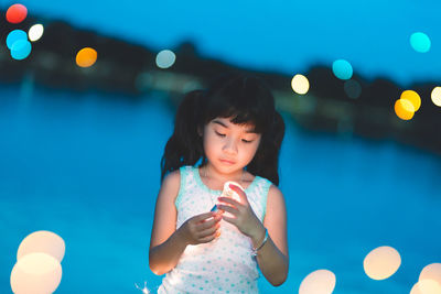 Girl looking at illuminated light bulb while standing against river at night