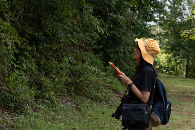 Botanist wearing hat looking at plants while holding book at forest