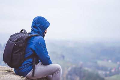 Rear view of man sitting on rock looking at landscape against sky