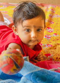 A cute indian baby holding a ball towards the camera while lying down on a bed-sheet over a mat.