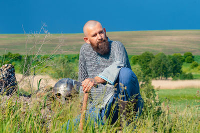 Viking. bald bearded man in metal chain mail over linen shirt sits on hill, leaning on ax. 