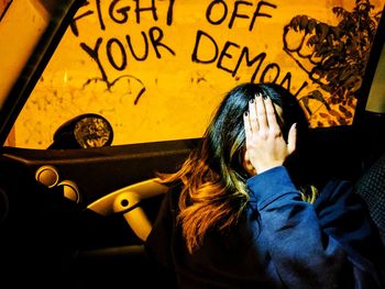 Woman covering face while sitting in car against graffiti on wall