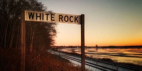Road sign by railroad tracks against clear sky during sunset