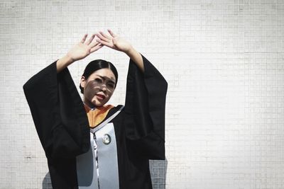 Portrait of young woman wearing graduation gown shielding eyes against wall