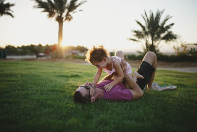 Father laying on grass holding his laughing daughter in sunset