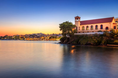 River by illuminated buildings against clear sky at sunset